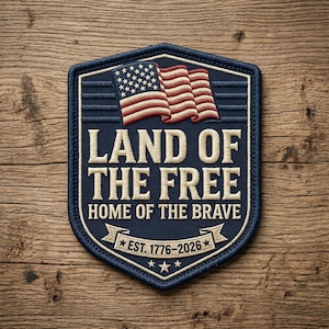 May include: A navy blue embroidered patch with the text "LAND OF THE FREE HOME OF THE BRAVE" and an American flag design. The patch includes the text "EST. 1776-2026" and is bordered with a cream-colored stitching.
