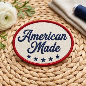 May include: Oval patch with the words "American Made" in navy blue script, surrounded by a red border. Five navy blue stars are below the text. The patch is on a woven surface.