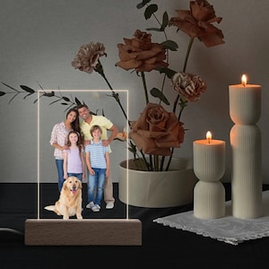 May include: A personalized LED photo frame featuring a family portrait with a golden retriever. The rectangular light-up panel sits on a wooden base. The background includes lit candles and a floral arrangement.