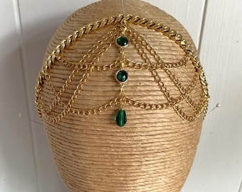 May include: Gold chain headpiece with three green gemstone accents. The chain is layered and has a teardrop-shaped gemstone dangling from the bottom.