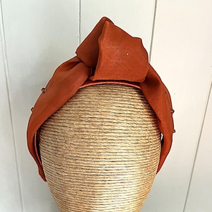 Puede incluir: Una diadema de tela naranja con la parte superior anudada, exhibida en una cabeza de maniquí. La diadema tiene una textura similar al satén y está adornada con pequeños adornos redondos. La cabeza del maniquí está cubierta con un material tejido.