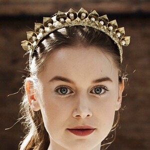 May include: A gold headband with a row of pearl-like beads and leaf-shaped accents. The headband is worn by a person with long blonde hair.