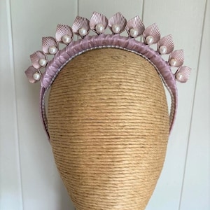 May include: A dusty pink headband featuring a series of upright, fan-shaped metallic petals arranged along the top. Each petal is accented with a small, round, white faux pearl at its base, and the band is wrapped in matching pink satin fabric.