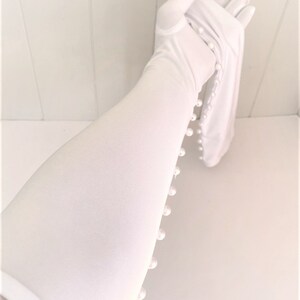 May include: White long gloves with pearl embellishments along the side. The gloves are made of a soft, stretchy fabric.