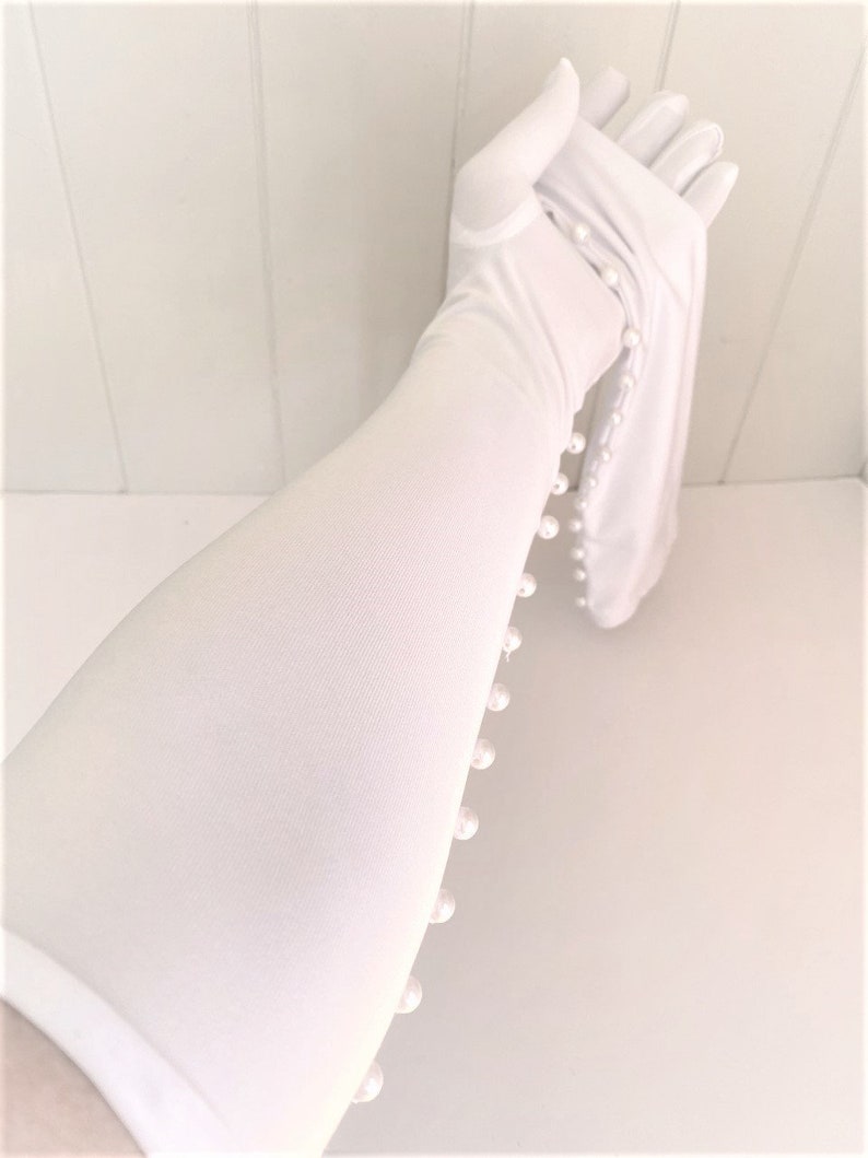 May include: White long gloves with pearl embellishments along the side. The gloves are made of a soft, stretchy fabric.