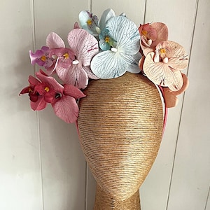 Könnte beinhalten: Ein floraler Haarschmuck mit Orchideenblüten in Rosa, Pfirsich und Blau. Die Blüten sind an einem rosa Haarband befestigt und auf einem Schaufensterkopf angeordnet. Ein dekoratives Accessoire.