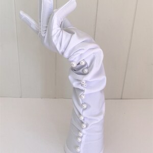 May include: White long gloves with pearl embellishments. The gloves are gathered at the wrist and have a smooth, satin-like finish.