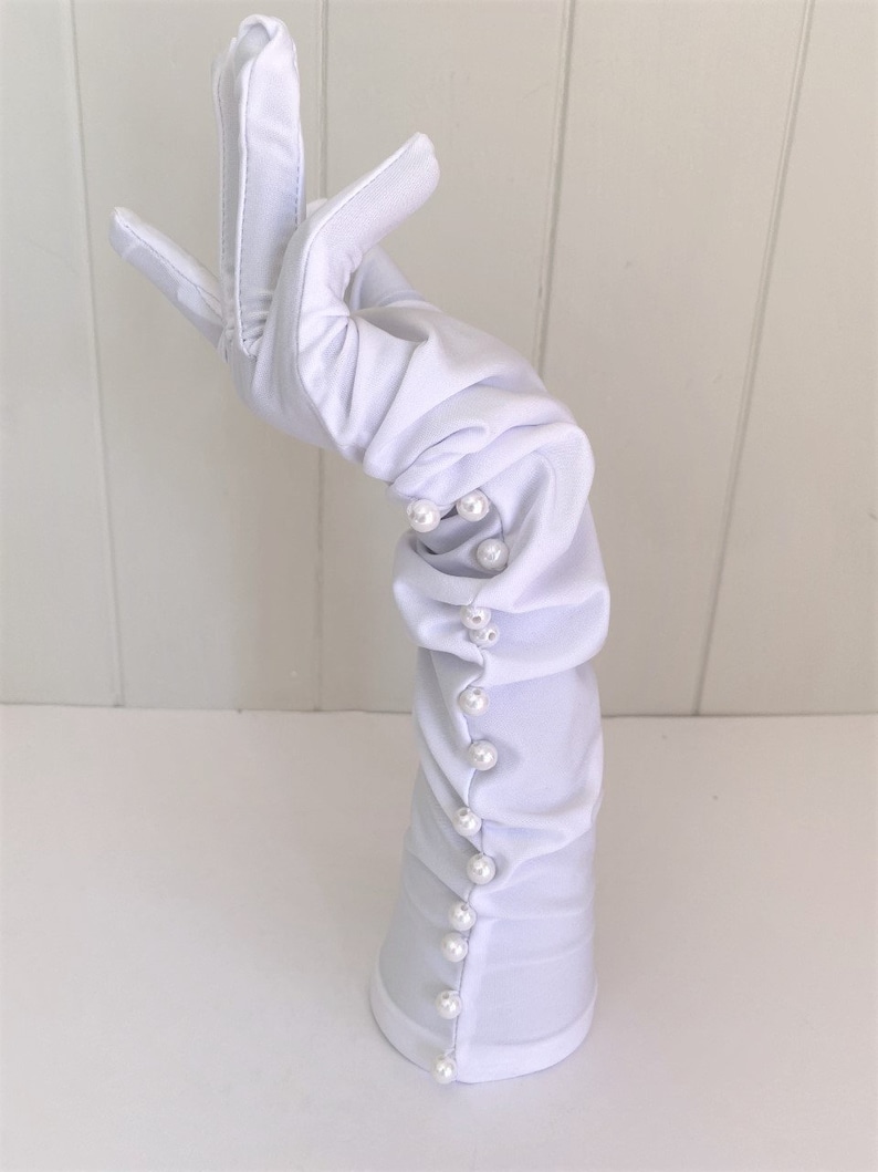 May include: White long gloves with pearl embellishments. The gloves are gathered at the wrist and have a smooth, satin-like finish.
