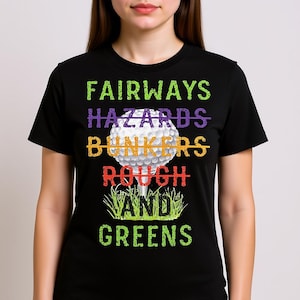 Puede incluir: Camiseta negra con un diseño temático de golf. El diseño presenta una pelota de golf sobre un tee, con las palabras "FAIRWAYS", "HAZARDS", "BUNKERS", "ROUGH" y "AND GREENS" en varios colores. El texto está tachado.