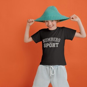 May include: A black t-shirt with the text "NUMBERS ARE MY SPORT" in white and gray lettering. The person is wearing a teal-colored hat and light blue pants against an orange background.