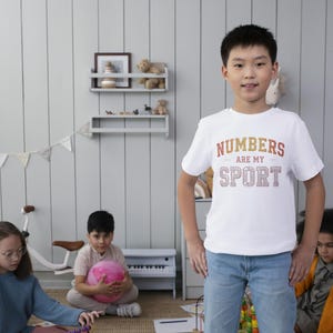 May include: A young person wearing a white t-shirt with the text "NUMBERS ARE MY SPORT" in orange and brown lettering. The person is wearing blue jeans and standing in a room with other children and toys.