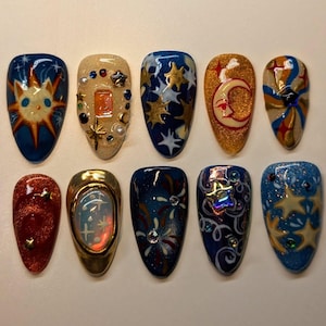 May include: A collection of ten artificial nails with celestial designs. Each nail features a different design, including stars, moons, and suns, in various colors like blue, gold, red, and beige. Some nails have glitter and embellishments.