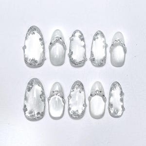 May include: A set of ten artificial nails in various oval shapes. The nails are primarily white with silver embellishments. Some nails feature silver floral designs, while others have a silver band with small, clear rhinestones.