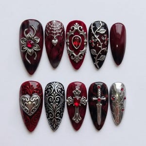 Può includere: Un set di dieci unghie finte a mandorla con una palette di colori bordeaux scuro e nero. I design includono ciondoli in metallo argentato come un fiore con una gemma rossa, una ragnatela con un cuore, cornici ornate, croci e un pugnale.