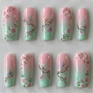 May include: A set of ten artificial nails with a gradient design. The nails transition from a soft pink at the top to a mint green at the bottom. Each nail features a hand-painted design of cherry blossom branches and butterflies.