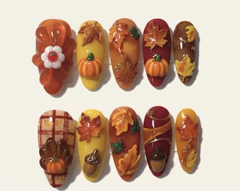 May include: A set of ten artificial nails with an autumn theme. Designs include pumpkins, fall leaves, acorns, and plaid patterns in shades of orange, yellow, and brown. Some nails feature 3D embellishments.