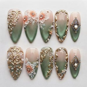 May include: A set of ten almond-shaped press-on nails featuring a soft nude to sage green gradient. The designs include gold filigree scrollwork, 3D peach-toned floral accents, small white pearls, and central diamond-shaped clear rhinestones.