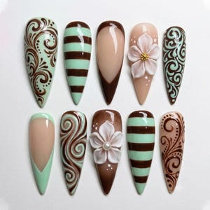 May include: A set of ten almond-shaped press-on nails featuring a variety of designs including mint green and brown swirls, horizontal brown and mint stripes, nude French tips with brown borders, and nude nails with 3D white floral accents.