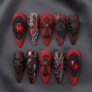 May include: A set of ten almond-shaped press-on nails in a deep red and black color palette. The designs feature ornate black frames with red faceted gems, intricate silver filigree, black crosses with red centers, cameos, and horned skull motifs.