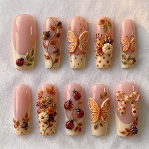May include: A set of ten press-on nails featuring a nude base with autumn-themed 3D embellishments including ladybirds, butterflies, pumpkins, acorns, pinecones, and a bow. The designs incorporate cream-coloured tips with polka dots and foliage.