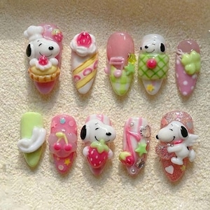 Puede incluir: Un conjunto de diez u&ntilde;as artificiales con dise&ntilde;os de personajes de dibujos animados en 3D. Las u&ntilde;as presentan a Snoopy, fresas, cupcakes y notas musicales en varios colores, incluyendo rosa, blanco, verde y amarillo. Las u&ntilde;as est&aacute;n decoradas con purpurina y otros adornos.