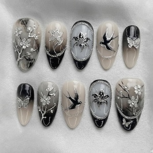 May include: A set of ten artificial nails with various designs. The nails feature a mix of colors, including black, white, and beige. Some nails have floral and butterfly embellishments, while others showcase bird and floral artwork.