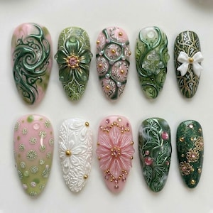 May include: A collection of ten intricately designed artificial nails. The nails feature various designs, including floral patterns, swirls, and embellishments with gold, pink, green, and white accents. Each nail showcases a unique, detailed design.