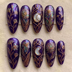 May include: Set of ten purple, almond-shaped press-on nails. Each nail features intricate gold-colored floral and celestial designs, including a crescent moon and various botanical elements. The nails have a glossy finish.