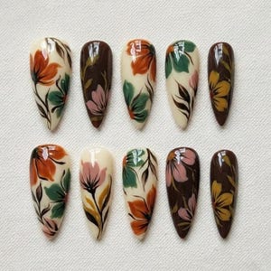 May include: A set of ten almond-shaped false nails. Each nail features a floral design in orange, green, pink, and yellow on a cream or brown base. The nails are arranged in two rows of five.