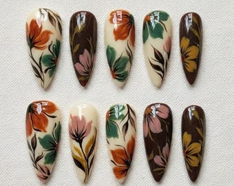 May include: A set of ten almond-shaped false nails. Each nail is painted with a floral design in shades of orange, green, pink, and yellow on a cream or brown base. The nails are arranged in two rows of five.