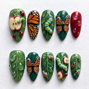 May include: A set of ten artificial nails with detailed, nature-themed designs. Each nail features a different design, including butterflies, caterpillars, apples, ladybugs, and water droplets, all set against a green leaf backdrop. The nails are oval-shaped and painted with vibrant colors.