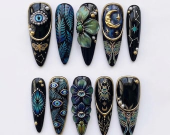 May include: A set of ten stiletto-shaped press-on nails featuring a black base with intricate designs including a blue eye, green beetle, gold crescent moon, fern leaves, constellations, and floral motifs accented with small white pearls and gold trim.