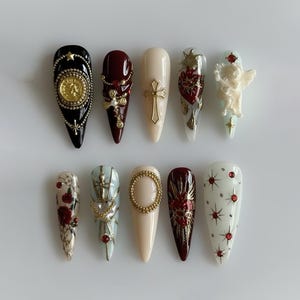 May include: Ten long, almond-shaped artificial nails with various designs. The nails feature gold crosses, coins, and heart motifs. The colour palette includes black, burgundy, cream, and white, with gold and red accents.