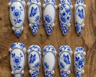 May include: A set of ten white and blue porcelain-style press-on nails. Each nail features a unique design, including floral patterns and butterfly motifs, framed with silver and accented with small pearls. The nails are arranged on a wooden surface.