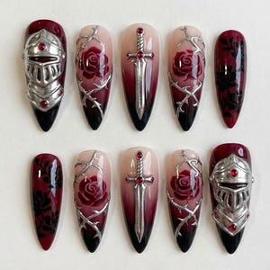 May include: A set of ten long, pointed false nails. Designs include a knight's helmet, roses, thorns, and swords. The nails feature a gradient from nude to black, with silver and red accents. The overall theme is gothic and romantic.