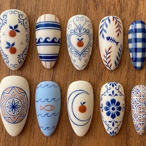 Könnte beinhalten: Zehn mandelförmige Kunstnägel mit verschiedenen Designs in Blau, Orange und Creme. Die Muster umfassen Blumen, Früchte und geometrische Formen, die an Porzellankunst erinnern. Auf einer Holzoberfläche angeordnet.