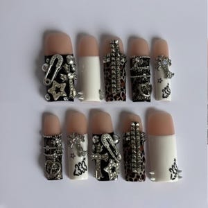 May include: A set of ten artificial nails with a nude base and white tips. Each nail is adorned with silver-colored embellishments, including crosses, stars, and safety pins. Some nails feature a leopard print design, and the number "233" is printed on some nails.