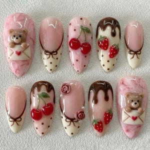 May include: A set of ten artificial nails with various designs. The nails feature pink, white, and brown colors, with decorative elements like cherries, strawberries, teddy bears, and bows. Some nails have a marble effect.