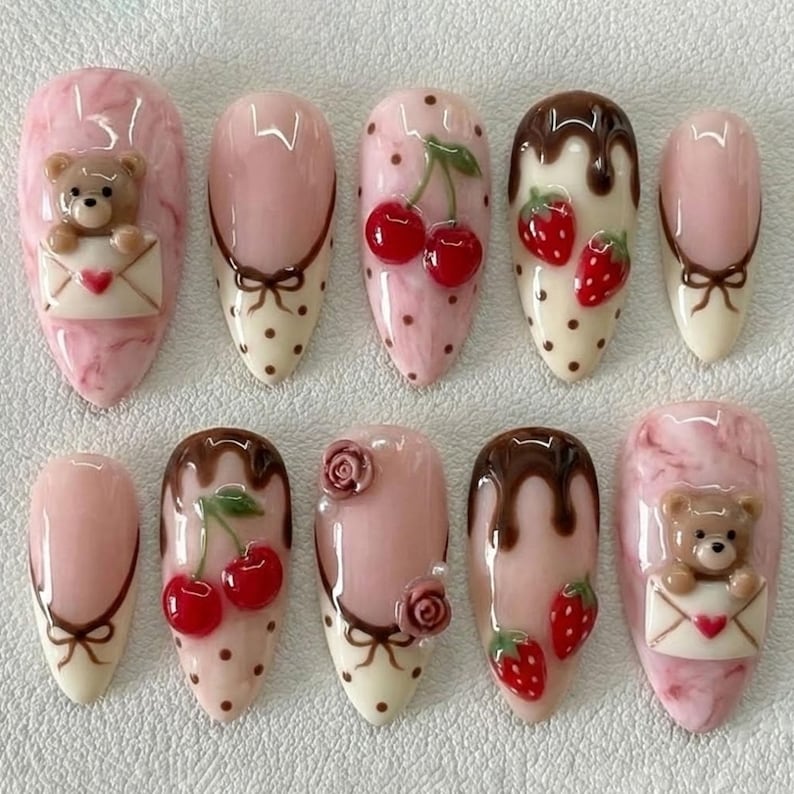 May include: A set of ten artificial nails with various designs. The nails feature pink, white, and brown colors, with decorative elements like cherries, strawberries, teddy bears, and bows. Some nails have a marble effect.