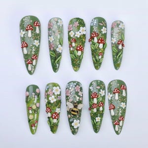 May include: A set of ten long, almond-shaped artificial nails. Each nail is painted a mossy green and decorated with miniature red-capped mushrooms, white flowers, and green leaves. One nail features a bumblebee.