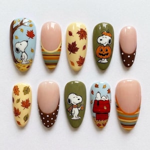 May include: A set of ten press-on nails featuring autumn-themed designs. The nails display illustrations of Snoopy, falling maple leaves, pumpkins, and a red doghouse. Other nails feature brown and white polka dots or horizontal stripes in fall colours.
