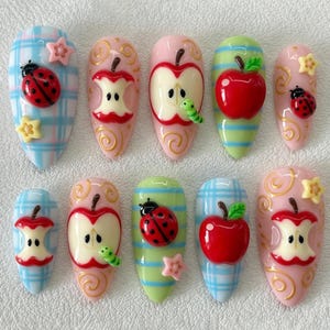 May include: A set of ten press-on nails featuring 3D designs of red ladybugs, whole red apples, apple cores, and small yellow stars. The nails have light blue plaid, light green striped, and solid pink backgrounds with gold swirl accents.