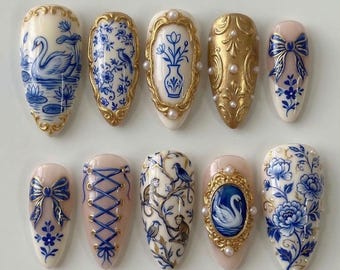 Puede incluir: Un conjunto de diez uñas artificiales con diseños intrincados. Las uñas presentan una variedad de patrones, incluyendo motivos florales azules y blancos, cisnes, lazos y adornos dorados. Algunas uñas tienen un diseño tipo corsé, mientras que otras muestran detalles de perlas.