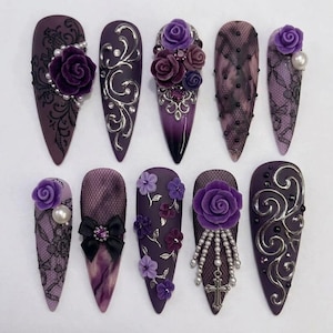 May include: A set of ten stiletto-shaped press-on nails in shades of purple and black. The designs feature 3D purple roses, silver filigree swirls, black lace patterns, pearl accents, a black bow with a central gem, and a silver cross charm.