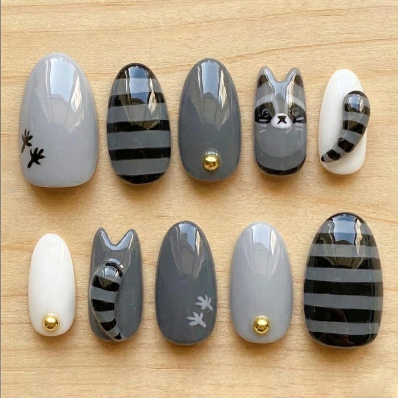 May include: A set of ten artificial nails with a raccoon theme. The nails are oval-shaped and feature designs in gray, black, and white. Some nails have raccoon faces, striped patterns, or paw prints. A gold stud accents some nails.