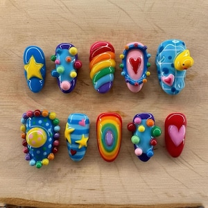 May include: A collection of ten colorful, artistic nail designs. Each nail features unique designs, including stars, hearts, rainbows, and abstract patterns. The nails are in various shades of blue, red, yellow, and green, with a glossy finish.
