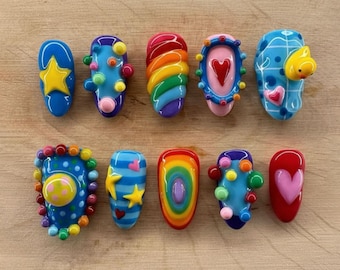 Op de afbeelding: Een verzameling van tien kleurrijke, artistieke nagelontwerpen. Elke nagel heeft unieke ontwerpen, waaronder sterren, harten, regenbogen en abstracte patronen. De nagels zijn in verschillende tinten blauw, rood, geel en groen, met een glanzende afwerking.