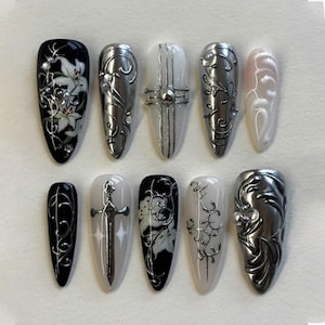 May include: A set of ten long, almond-shaped artificial nails. Designs include silver metallic embellishments, floral patterns, and a sword. Colors include black, white, silver, and a pale pink. The nails are arranged on a white surface.
