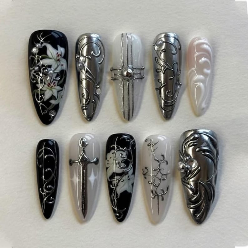 May include: A set of ten long, almond-shaped artificial nails. Designs include silver metallic embellishments, floral patterns, and a sword. Colors include black, white, silver, and a pale pink. The nails are arranged on a white surface.