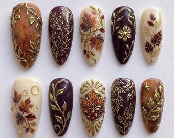 May include: A set of ten almond-shaped press-on nails featuring autumn-inspired designs. The collection includes cream, deep plum, and textured brown bases adorned with raised gold metallic leaf patterns, painted maple leaves, and clear water droplets.